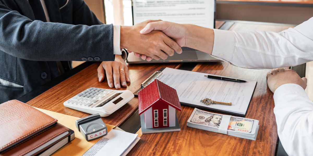 Two people shaking hands over a desk with a small house model, cash, documents, and a calculator